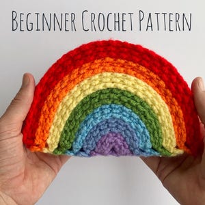 Op de afbeelding: Een regenboog haakpatroon, met rood, oranje, geel, groen, blauw en paars garen, wordt in twee handen gehouden. De tekst "BEGINNER CROCHET PATTERN" staat bovenaan de afbeelding.