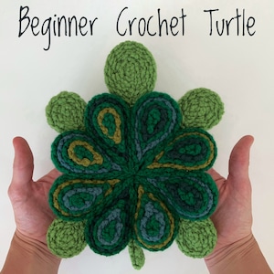 May include: A green crocheted turtle with a multi-coloured shell. The turtle is made with different shades of green, blue, and yellow yarn. The turtle is being held in the hands of a person.