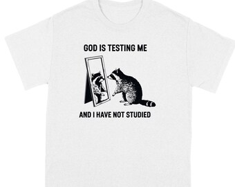 May include: White t-shirt with a black and white illustration of a raccoon looking in a mirror. The text "God is testing me and I have not studied" is printed on the shirt.