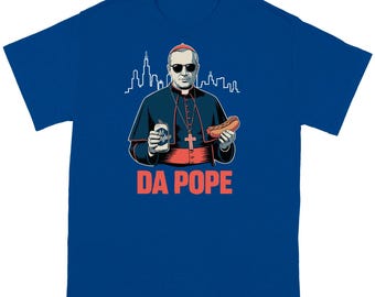May include: Royal blue t-shirt featuring a cartoon illustration of a figure in religious garb wearing sunglasses, holding a can of beer and a hot dog. The text "DA POPE" is printed in red below the image, with a city skyline in the background.