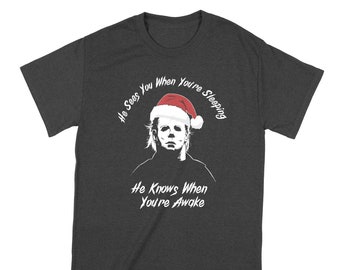 May include: A dark heather grey t-shirt with a white graphic of Michael Myers wearing a Santa hat. The text "He Sees You When You're Sleeping He Knows When You're Awake" is printed on the shirt.