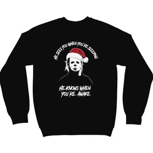 Könnte beinhalten: Schwarzer Pullover mit einem weißen Aufdruck eines Mannes mit einem Weihnachtsmann-Hut. Der Text lautet "Er sieht dich, wenn du schläfst. Er weiß, wenn du wach bist."
