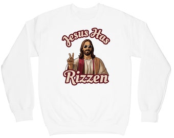 May include: White sweatshirt with a graphic of Jesus wearing sunglasses and making a peace sign. The text "Jesus Has Rizzen" is printed in red below the image.