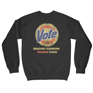 May include: Black sweatshirt with a vintage-style graphic of a cleaning product logo that says "Vote" in blue text. The logo is surrounded by a red and yellow circle. Below the logo, the text "Removes Stubborn Orange Stains" is printed in white.