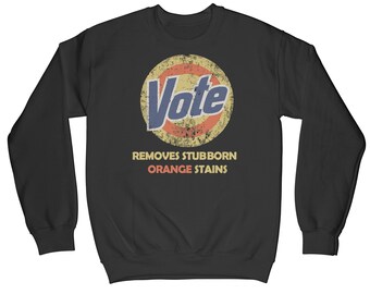 May include: Black sweatshirt with a vintage-style graphic of a cleaning product logo that says "Vote" in blue text. The logo is surrounded by a red and yellow circle. Below the logo, the text "Removes Stubborn Orange Stains" is printed in white.