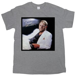 以下が含まれることがあります： 白いスーツを着た男性の白黒画像がプリントされたグレーのTシャツ。画像は「Phil Kessel Thriller」というテキストが書かれたレコードアルバムのジャケットです。