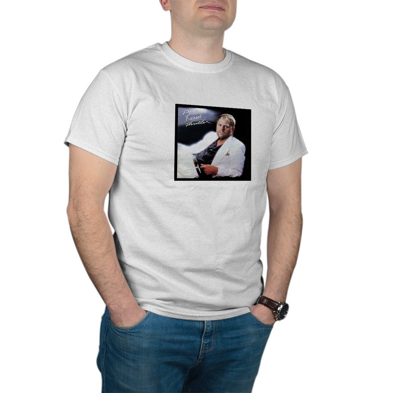 以下が含まれることがあります： 白いTシャツに、白の スーツを着た男性の白黒写真がプリントされています。写真は、アルバムのジャケットで、そこに「The Rebel Within」という文字が書かれています。
