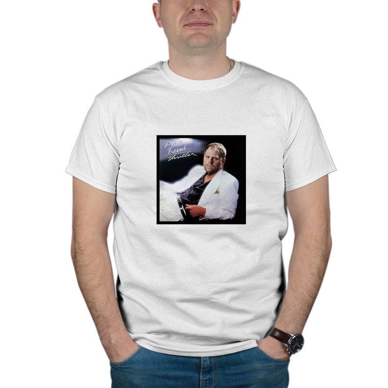 以下が含まれることがあります： 前面にフィル・ケッセルの写真がプリントされた白いTシャツ。写真は彼の「スリラー」アルバムのジャケットから。写真には、白いスーツを着て椅子に座り、マイクを持ったケッセルが写っている。