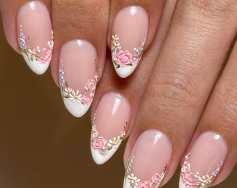 May include: Almond-shaped nails featuring a classic French manicure with a nude base and white tips. Each nail is decorated with delicate 3D floral art, including small pink roses, white daisies, tiny blue sprigs, and small white pearls.