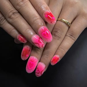 Può includere: Unghie ovali rosa e rosse con disegni a cuore e croce. Le unghie hanno una finitura lucida e sono decorate con cuori rosa neon e motivi a croce rossi. Un anello d'oro è visibile su un dito.