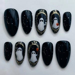 May include: A set of ten almond-shaped press-on nails with a glossy black base featuring white speckles. Several nails display hand-painted white ghosts framed in metallic gold, with one ghost holding an orange balloon and another a pumpkin.