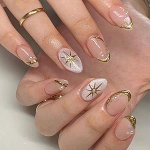 Peut inclure: Ongles de forme ovale avec une base nude, des accents métalliques dorés, de petites perles blanches et des motifs d'étoiles à paillettes argentées. La manucure comprend des bagues dorées sur les doigts, soulignant un nail art détaillé.