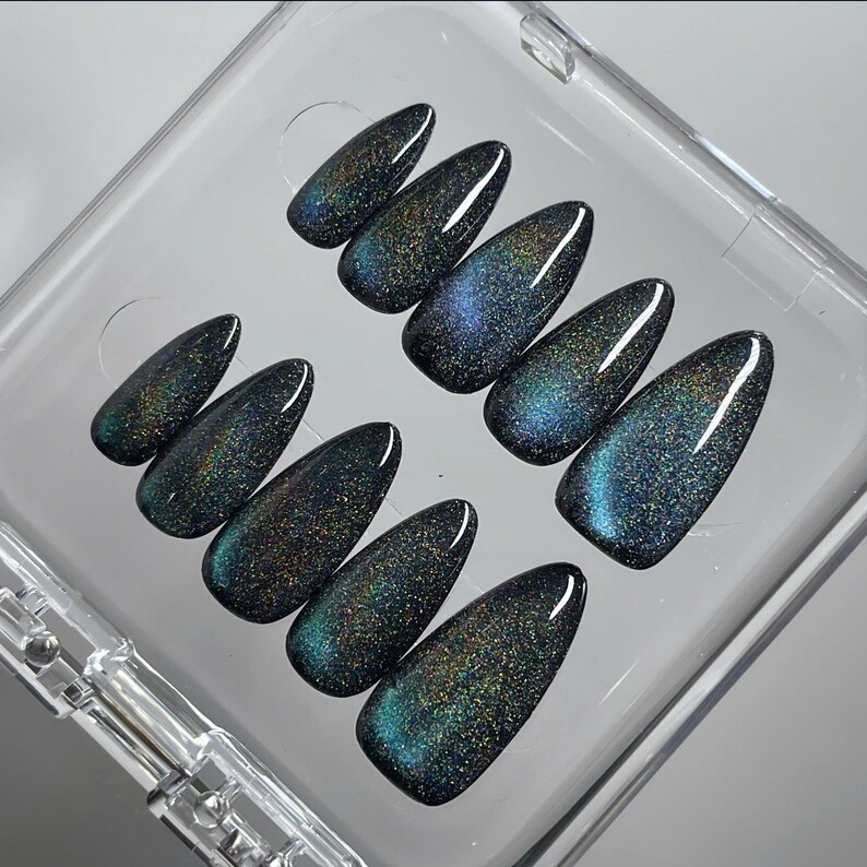 May include: A set of ten almond-shaped press-on nails featuring a dark, magnetic cat-eye finish. The surface displays a deep teal and black base with shimmering, holographic glitter particles that create a reflective, multi-dimensional effect.