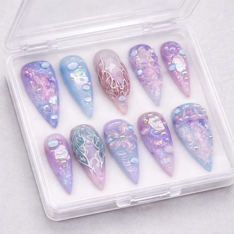May include: A set of ten stiletto-shaped press-on nails in a clear plastic storage case. The nails feature a color palette of pastel blue and purple, decorated with iridescent flakes, small round beads, and intricate white line art designs.
