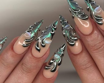 Può includere: Unghie artificiali lunghe a stiletto con una base color nude. Ogni unghia è decorata con vortici e accenti metallici argentati in 3D in rilievo, sovrapposti a uno smalto magnetico effetto occhio di gatto dai riflessi verdi e blu.