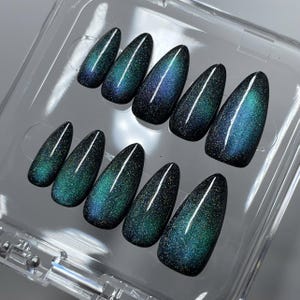 May include: A set of ten almond-shaped press-on nails featuring a dark base with a shimmering, magnetic cat-eye effect in shades of deep teal, emerald green, and midnight blue, arranged in two rows inside a clear plastic storage container.