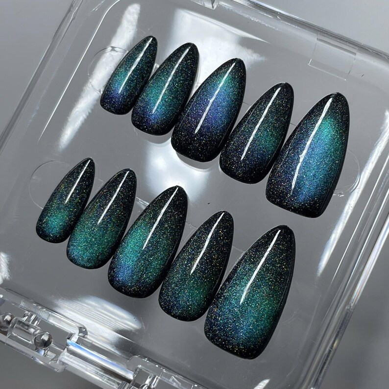 May include: A set of ten almond-shaped press-on nails featuring a dark base with a shimmering, magnetic cat-eye effect in shades of deep teal, emerald green, and midnight blue, arranged in two rows inside a clear plastic storage container.