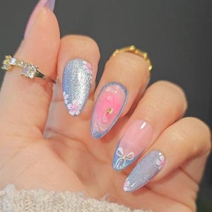 May include: A hand with long, almond-shaped nails. The nails feature a variety of designs, including glitter, floral patterns, and pink and blue swirls. Some nails have small bow accents and gold embellishments. The nails are painted in shades of pink, blue, and silver.