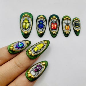 May include: Set of ten green and gold oval-shaped fake nails with different colored beetle designs. Each nail has a gold rim and gold beads around the base.