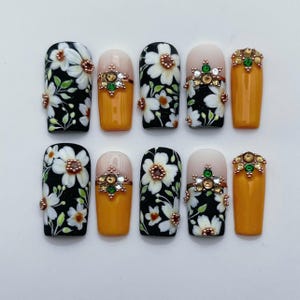 May include: A set of ten press-on nails featuring a mix of designs. Some nails have a black base with white floral patterns and small metallic beads, while others feature a solid mustard yellow color with decorative green and clear rhinestones.
