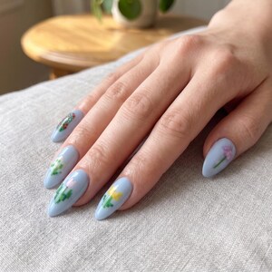 Puede incluir: U&ntilde;as en forma de almendra pintadas con esmalte azul b&iacute;garo claro. Cada u&ntilde;a presenta un peque&ntilde;o y distintivo dise&ntilde;o de arte de p&iacute;xeles con diversas flores y vegetaci&oacute;n coloridas, incluyendo motivos florales amarillos, rosas y verdes con acabado liso.