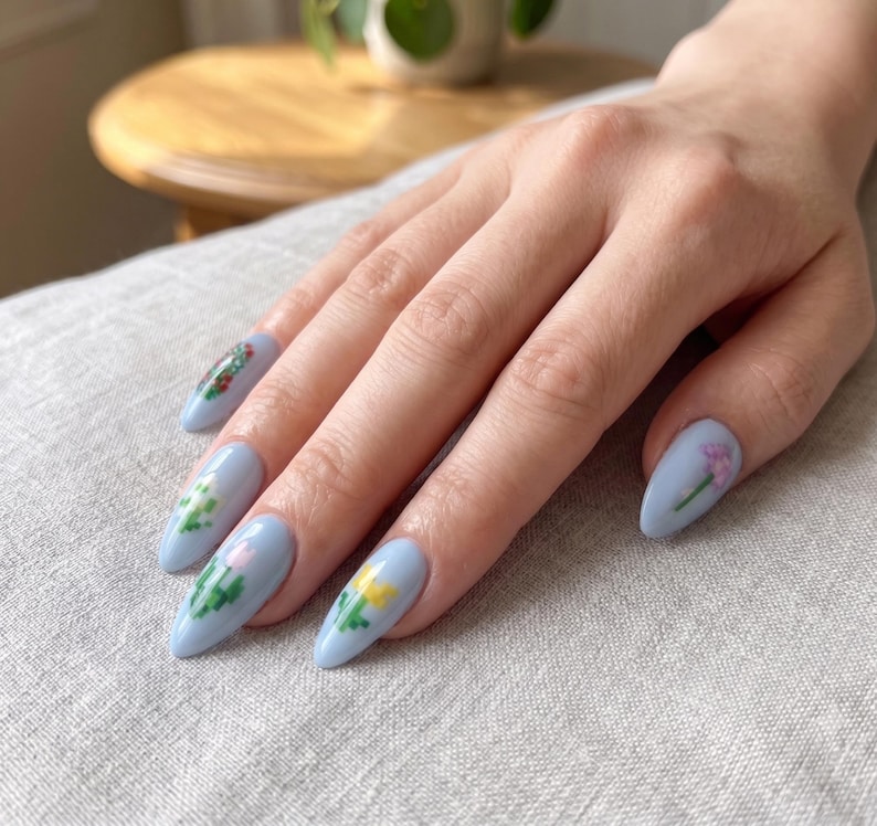 Puede incluir: U&ntilde;as en forma de almendra pintadas con esmalte azul b&iacute;garo claro. Cada u&ntilde;a presenta un peque&ntilde;o y distintivo dise&ntilde;o de arte de p&iacute;xeles con diversas flores y vegetaci&oacute;n coloridas, incluyendo motivos florales amarillos, rosas y verdes con acabado liso.
