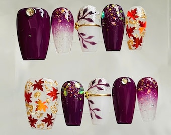 May include: A set of ten press-on nails featuring a variety of designs including solid deep purple with a single rhinestone, purple to white ombre with glitter, white with purple leaf accents and gold lines, purple with multicolored glitter, and white with red maple leaves.