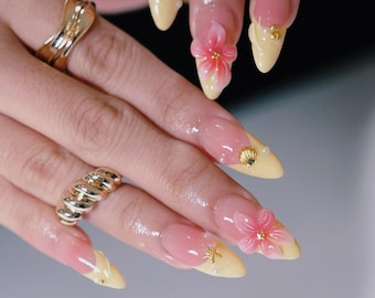 Puede incluir: Uñas en forma de almendra con puntas francesas de color amarillo pálido, decoradas con flores rosas en relieve, dijes metálicos dorados de estrellas de mar y conchas, y pequeñas perlas blancas, junto a dos anillos de metal dorado.