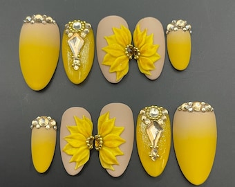 May include: Set of ten false nails with a yellow and beige ombre design. Some nails feature a 3D sunflower design with rhinestones, while others have a single rhinestone design.