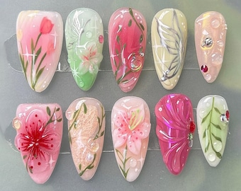 May include: A set of ten artificial nails, each with a unique floral design. The nails feature a variety of colors, including pink, green, and white, with intricate details like butterflies and water droplets. The nail shapes vary.