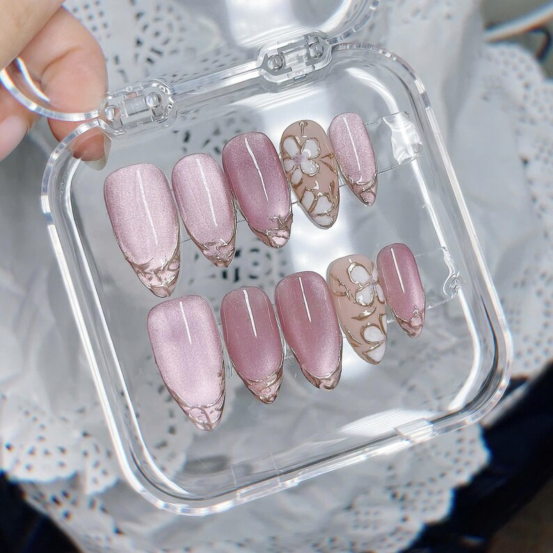 Op de afbeelding: Een set van tien kunstnagels in een doorzichtig plastic doosje. De nagels hebben een glinsterende mauve kattenoog-afwerking, waarbij twee nagels een wit bloemmotief op een nude basis hebben. Elke nagel heeft een afgeronde amandelvorm.
