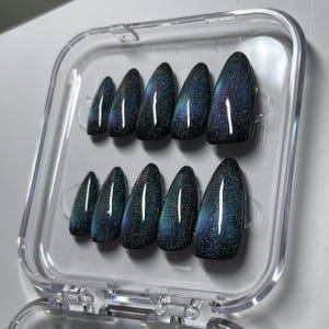 May include: A set of ten almond-shaped press-on nails arranged in a clear plastic storage case. Each nail features a dark, magnetic cat-eye finish with shimmering particles that create a deep, multi-dimensional effect in shades of blue and green.