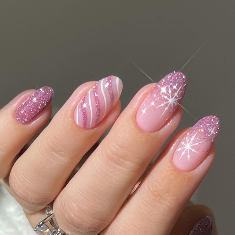 May include: Oval-shaped fingernails featuring a festive manicure with light pink polish, sparkling pink glitter accents, white diagonal stripes, and delicate white snowflake designs on the tips, creating a textured and shimmering appearance.