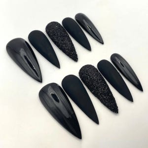 May include: A set of ten stiletto-shaped press-on nails in black. The collection features a mix of finishes, including glossy black, matte black, and textured black glitter nails, arranged in two rows of five on a plain white surface.