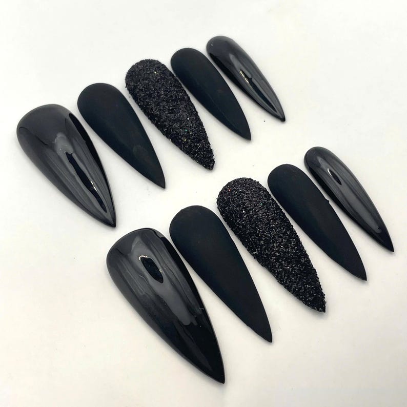 May include: A set of ten stiletto-shaped press-on nails in black. The collection features a mix of finishes, including glossy black, matte black, and textured black glitter nails, arranged in two rows of five on a plain white surface.