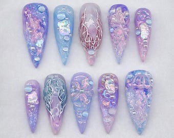 May include: A set of ten press-on nails in varying sizes, featuring a color palette of lavender, blue, and purple. The designs include iridescent flakes, small silver beads, white line art, and three-dimensional textures resembling jellyfish and floral patterns.