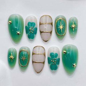 May include: A set of ten press-on nails featuring a gradient of green and translucent white. Designs include gold star charms, small gold spheres, painted green four-leaf clover shapes, and raised gold metallic lines over the nail surface.