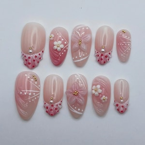 May include: A set of ten almond-shaped press-on nails featuring a translucent pink base. The designs include white floral accents, a butterfly motif, white dotted lines, and small gold and white beads. Some nails have a pink gradient tip with dots.