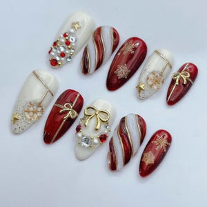 May include: A set of ten almond-shaped press-on nails featuring a holiday theme with red, white, and gold colors. Designs include candy cane stripes, gold snowflake decals, pearl and rhinestone accents, and raised gold bow charms on a glossy finish.