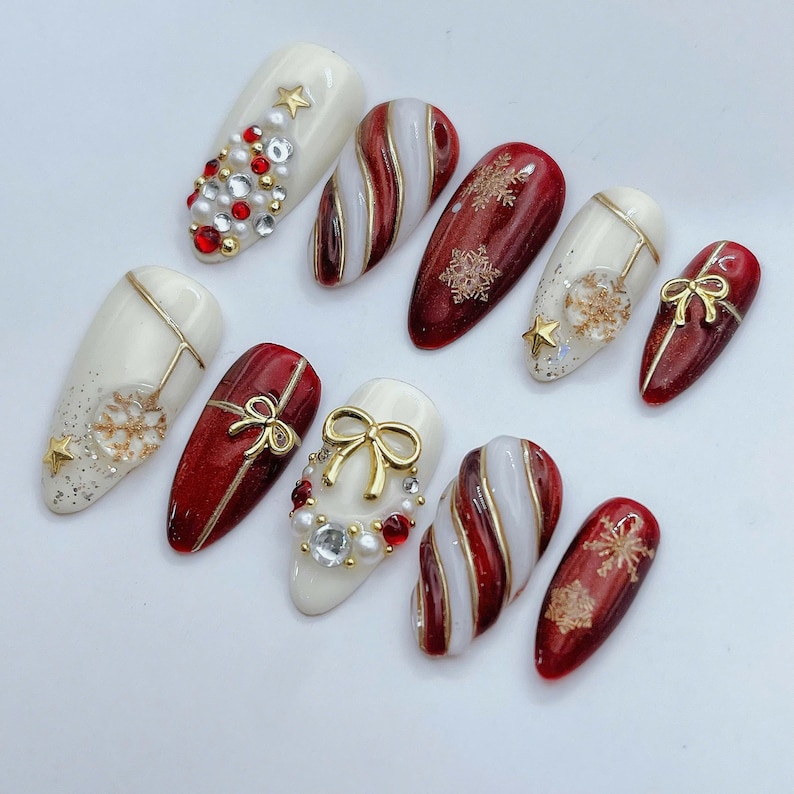 May include: A set of ten almond-shaped press-on nails featuring a holiday theme with red, white, and gold colors. Designs include candy cane stripes, gold snowflake decals, pearl and rhinestone accents, and raised gold bow charms on a glossy finish.