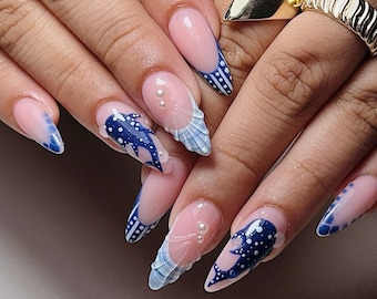 Puede incluir: Uñas largas en forma de almendra con base nude, patrones de tiburón ballena en azul y blanco, puntas francesas con puntos blancos y texturas de concha en 3D. Perlas blancas decoran la base, junto con dos anillos de color dorado.