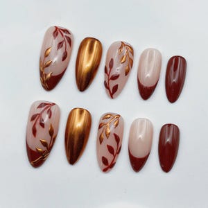 May include: A set of ten press-on nails featuring a mix of designs including solid metallic copper, solid deep burgundy, burgundy French tips on a nude base, and nude nails with painted metallic copper and burgundy leaf vine patterns.