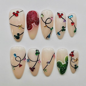 May include: A set of ten almond-shaped press-on nails with a nude base. Eight nails feature black hand-painted lines resembling string lights with colourful gem accents. Two nails feature glittery Mickey Mouse head shapes in red and green.