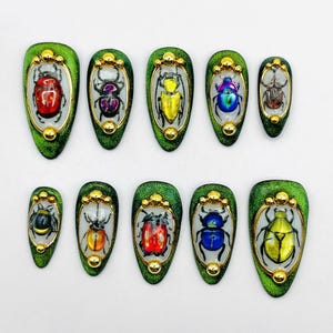 Peut inclure: Dix autocollants pour ongles de forme ovale, de couleur verte avec une bordure dorée et des motifs de scarabées colorés.