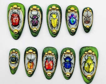 May include: A set of ten press-on nails featuring a dark green glitter finish. Each nail displays a unique beetle illustration encased in a gold-toned oval frame with small gold spheres at the top and bottom, showcasing various colors like red, blue, yellow, and brown.