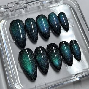 May include: A set of ten almond-shaped press-on nails featuring a dark, magnetic cat-eye finish. The nails display a deep black base color with a shimmering, iridescent teal and blue gradient effect that shifts across the surface of each nail.