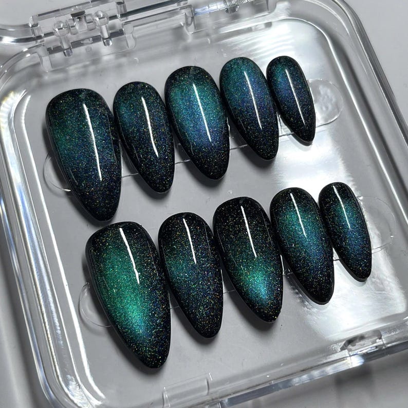 May include: A set of ten almond-shaped press-on nails featuring a dark, magnetic cat-eye finish. The nails display a deep black base color with a shimmering, iridescent teal and blue gradient effect that shifts across the surface of each nail.