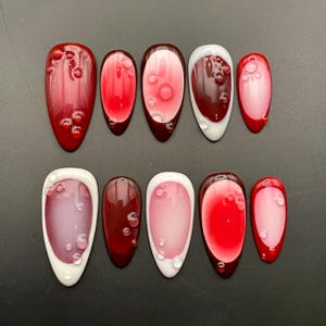 Può includere: Dieci unghie finte ovali di colore rosso e bianco con un design testurizzato a forma di bolle. Le unghie sono disposte in due file da cinque.