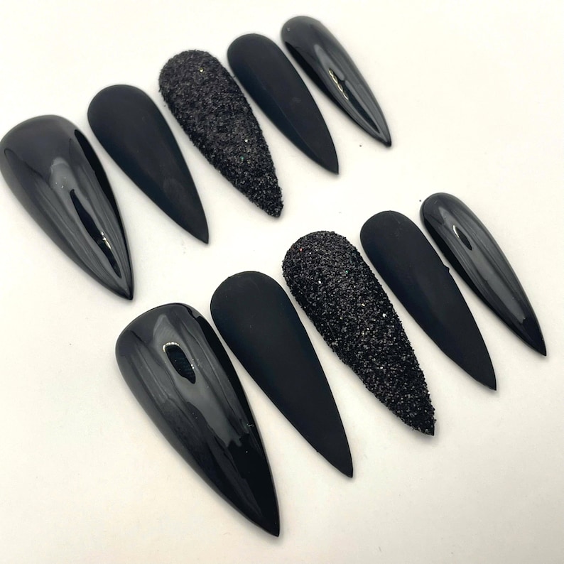 May include: A set of ten stiletto-shaped press-on nails in black. The collection features a mix of finishes including glossy black, matte black, and textured black glitter nails, all arranged in two rows of five on a plain white surface.