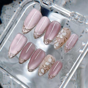 Op de afbeelding: Een set van tien kunstnagels in een doorzichtig plastic doosje. De nagels hebben een mauve kattenoog-afwerking met metallic gouden lijnaccenten. Sommige nagels zijn effen mauve, terwijl andere zijn voorzien van witte bloemmotieven.